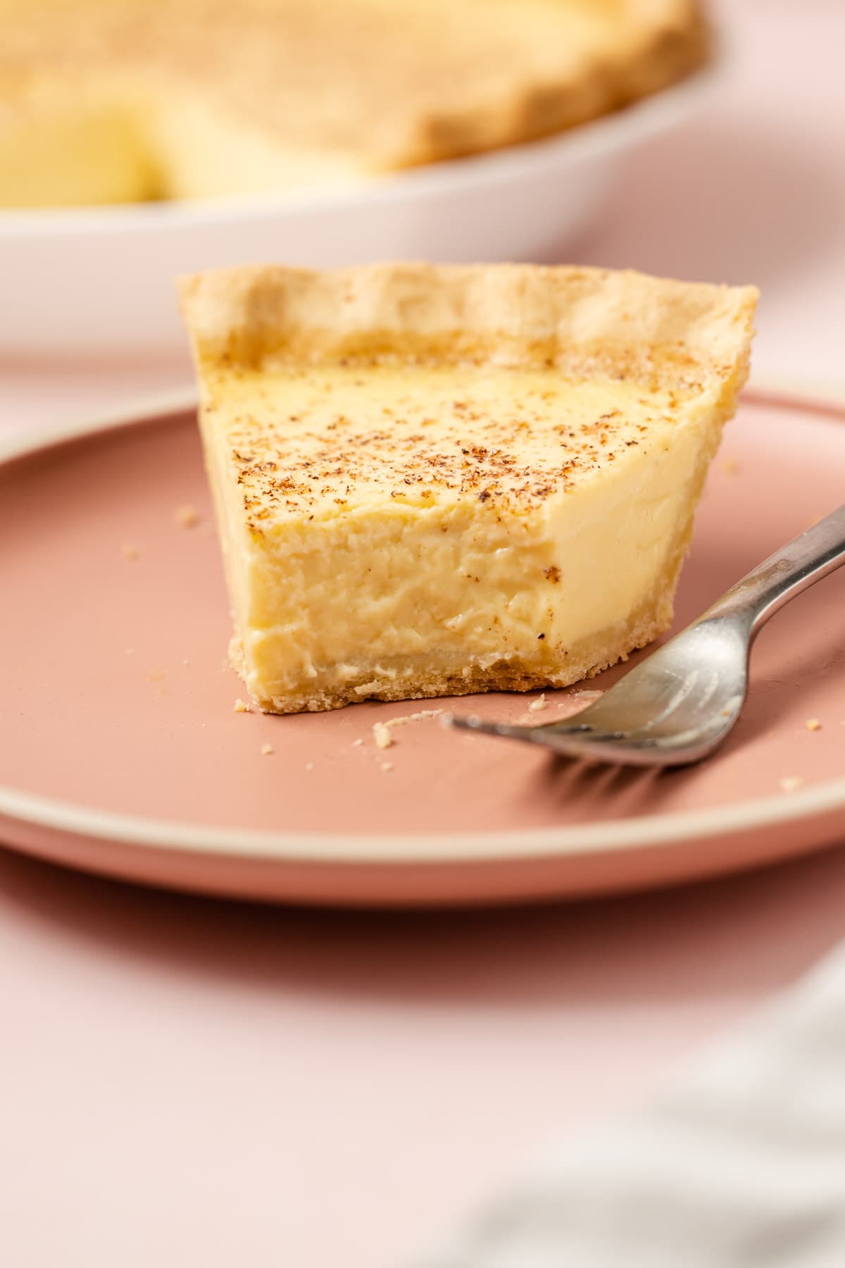 A slice of classic custard pie on a pink plate with a few bites taken from it.