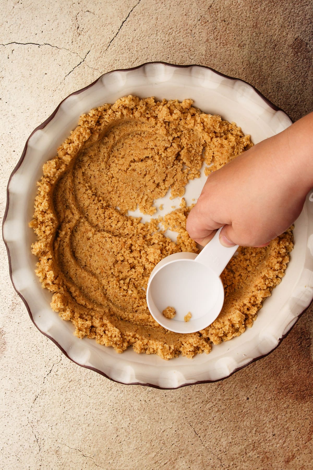 Pushing crumb crust ingredients into shape using a measuring cup in a pie dish.
