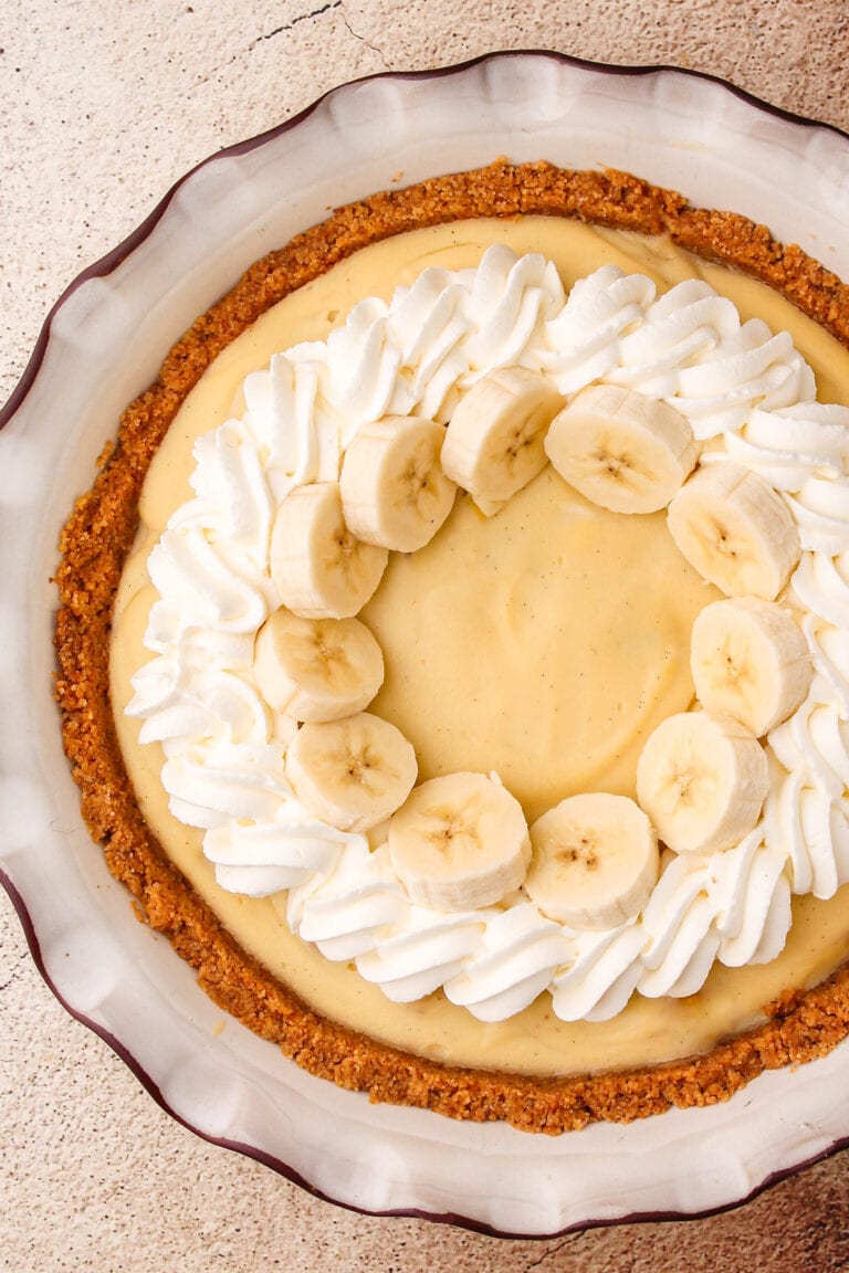 A fully assembled and garnished banana cream pie with a crumb crust, piped whipped cream and sliced fresh bananas.