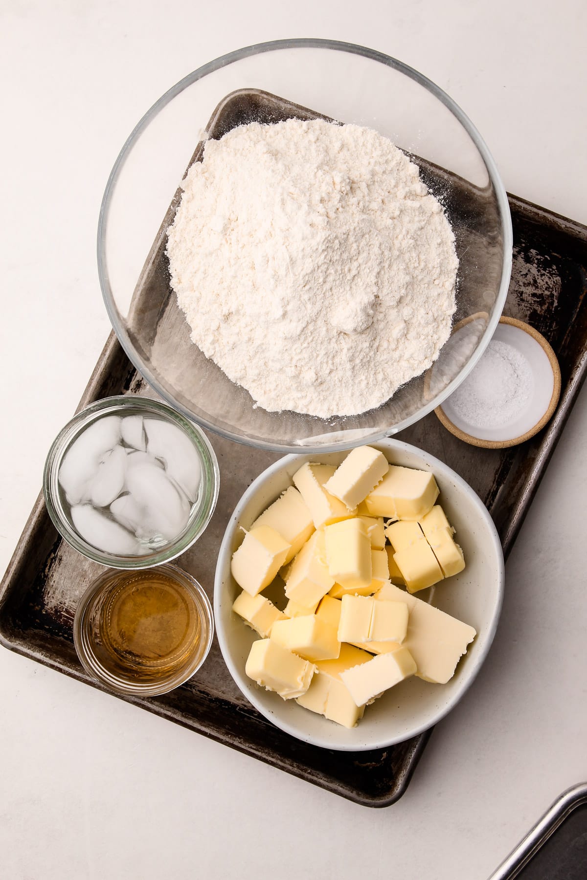 A baking sheet with bowls of ingredients on it, including flour, cubes of butter, cornstarch, salt, apple cider vinegar, and ice water