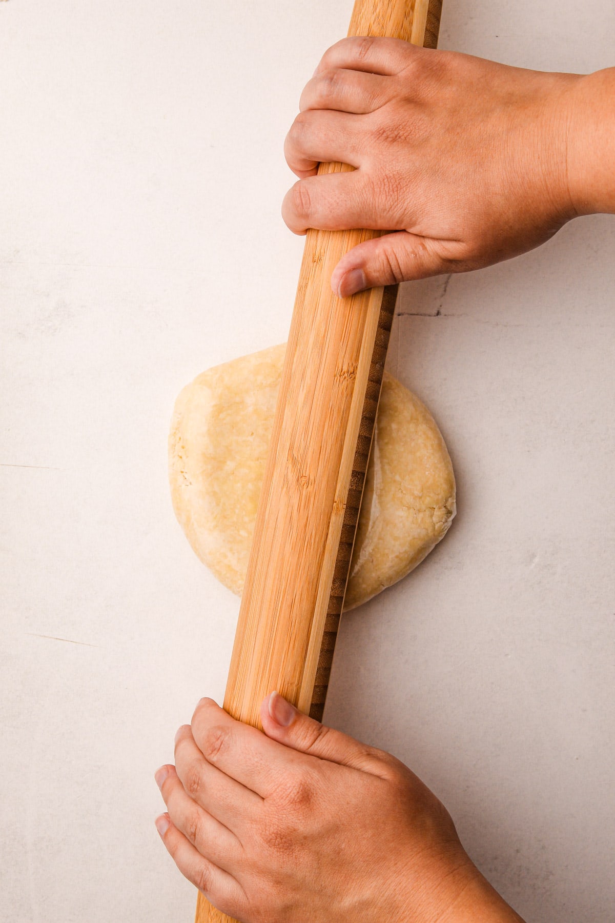 Two hands using a wooden rolling pin to flatten a disc of plastic-wrapped pie dough