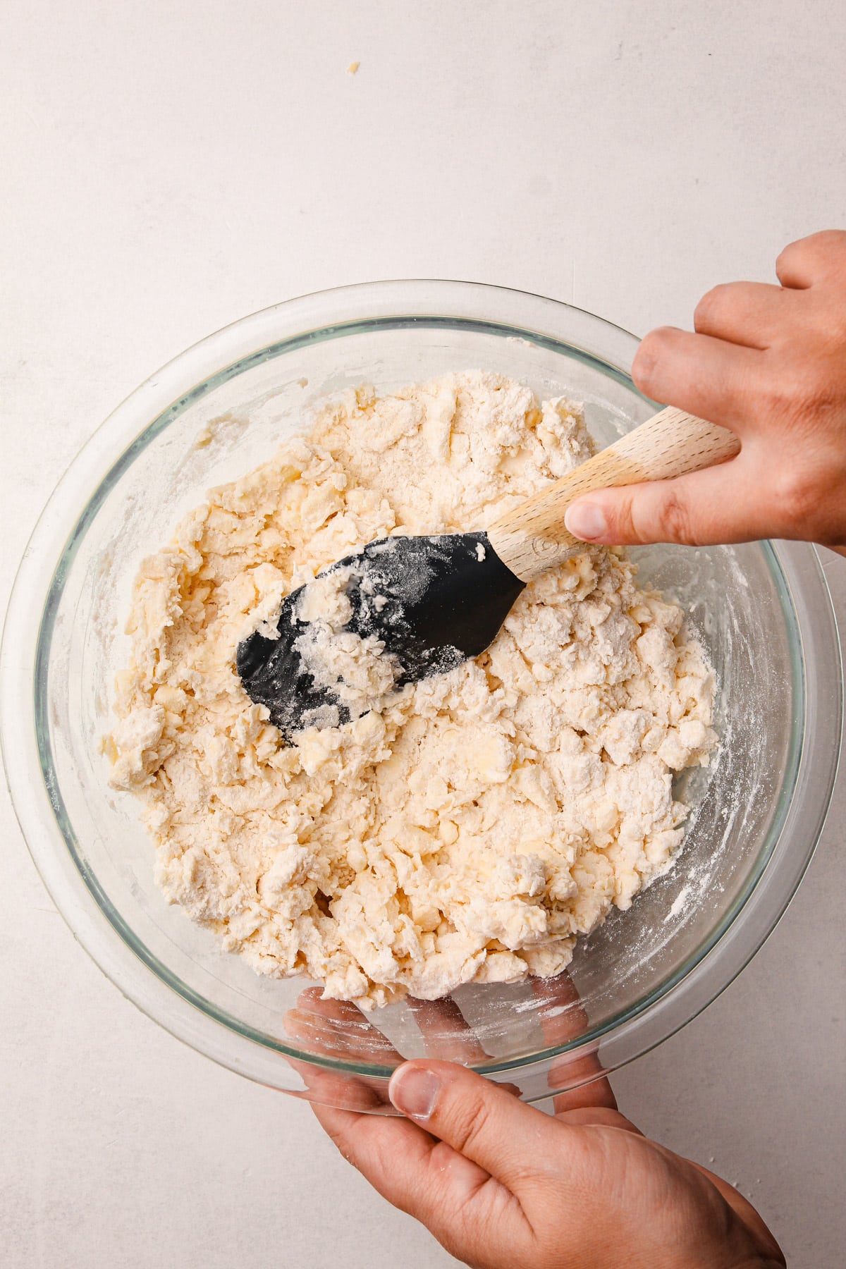 A hand using a rubber spatula to mix together butter pieces and a flour mixture in a glass bowl