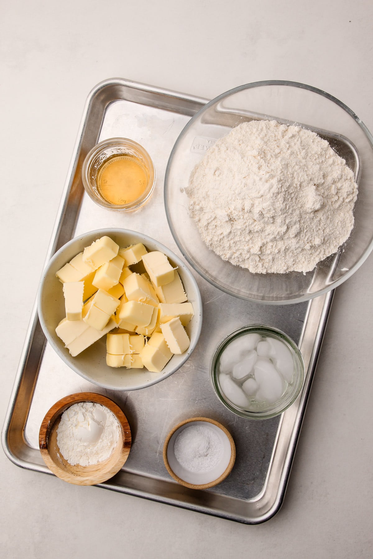 A baking sheet with bowls of ingredients on it, including flour, cubes of butter, cornstarch, salt, apple cider vinegar, and ice water