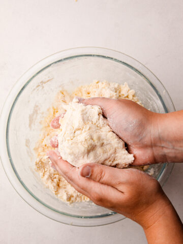 Two hands holding a ball of pie dough over a bowl with more flour and butter in it