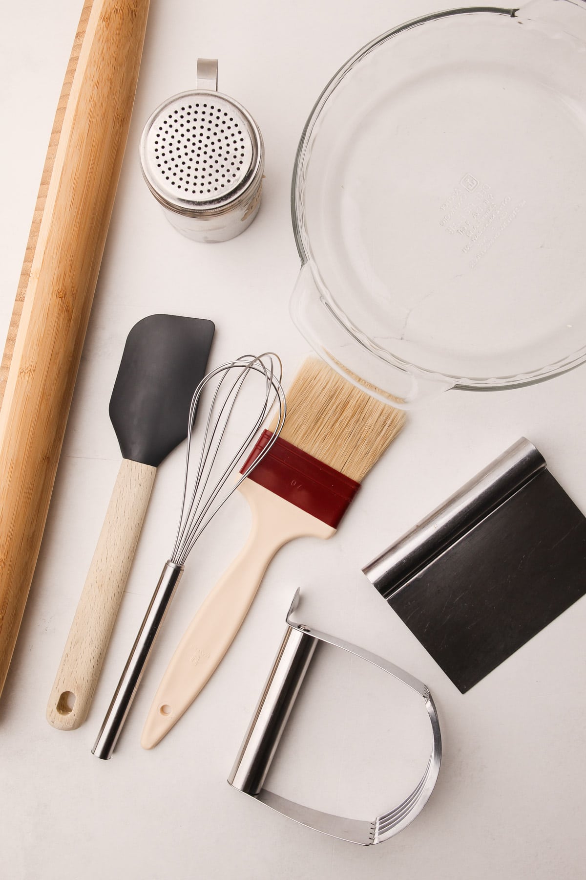 A wooden rolling pin, glass pie plate, rubber spatula, whisk, pastry brush, bench scraper, flour shaker, and handheld pastry blender on a counter surface
