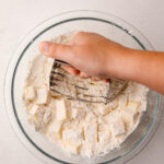 A hand using a pastry blender to mash butter into flour in a glass bowl