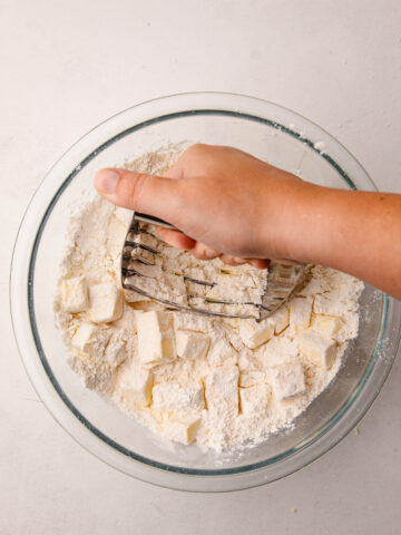 A hand using a pastry blender to mash butter into flour in a glass bowl