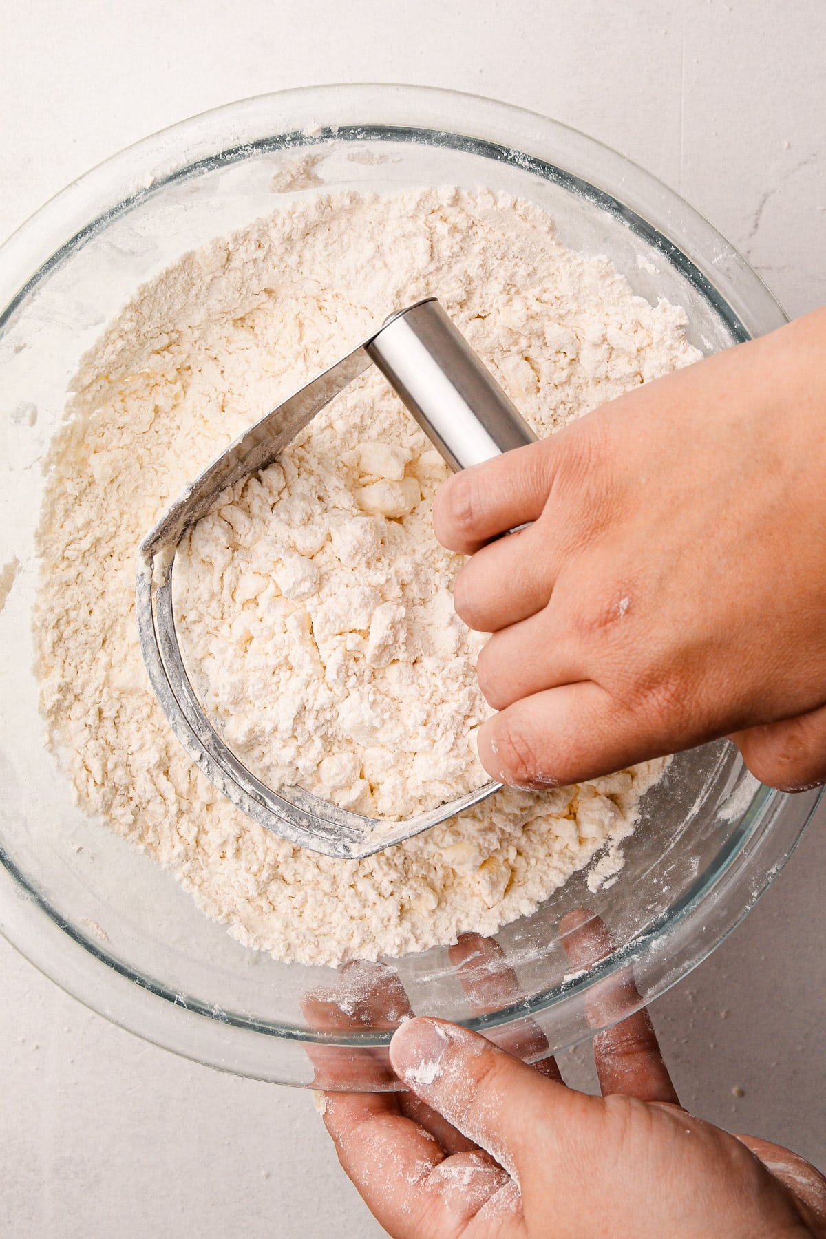 One hand using a pastry blender to mix together flour and butter in a bowl, with a second hand holding the bowl on the side