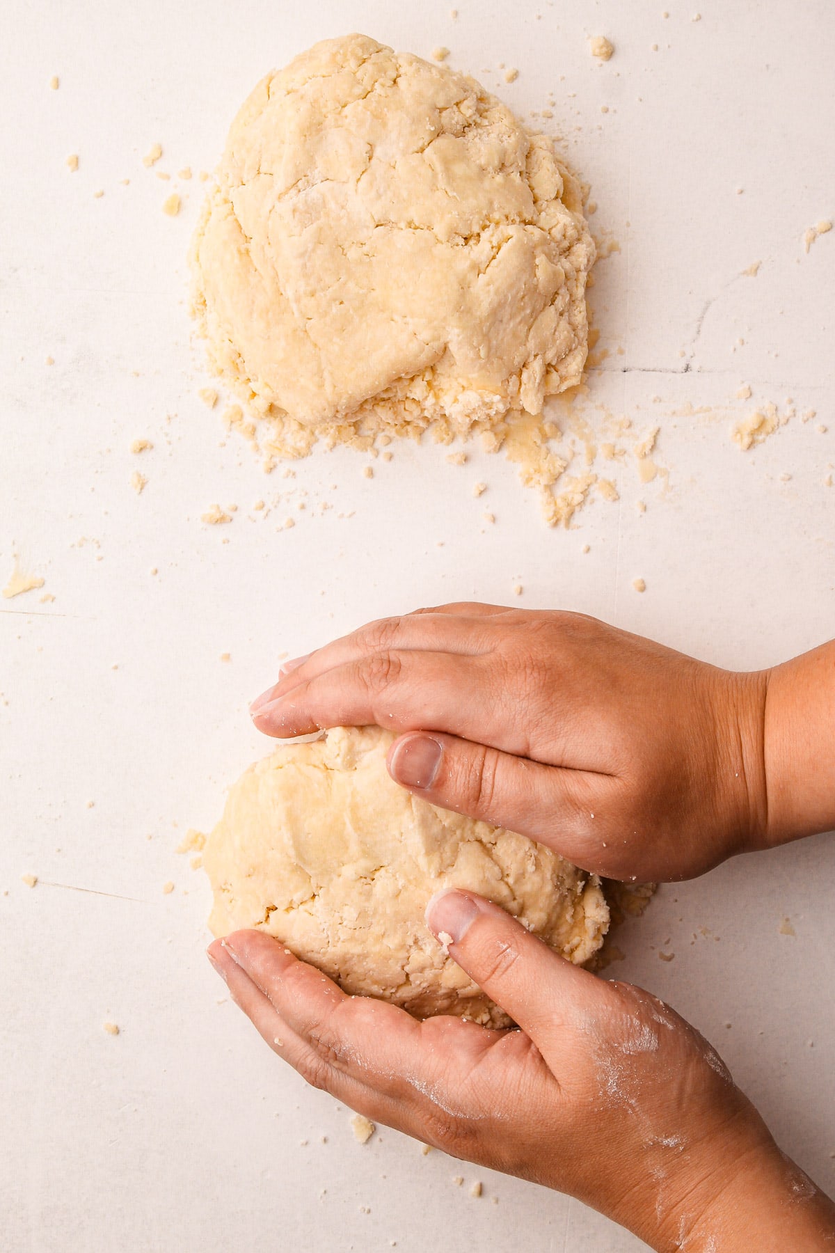 Two hands pressing together a mound of pie dough on a counter surface, with a second mound of dough above
