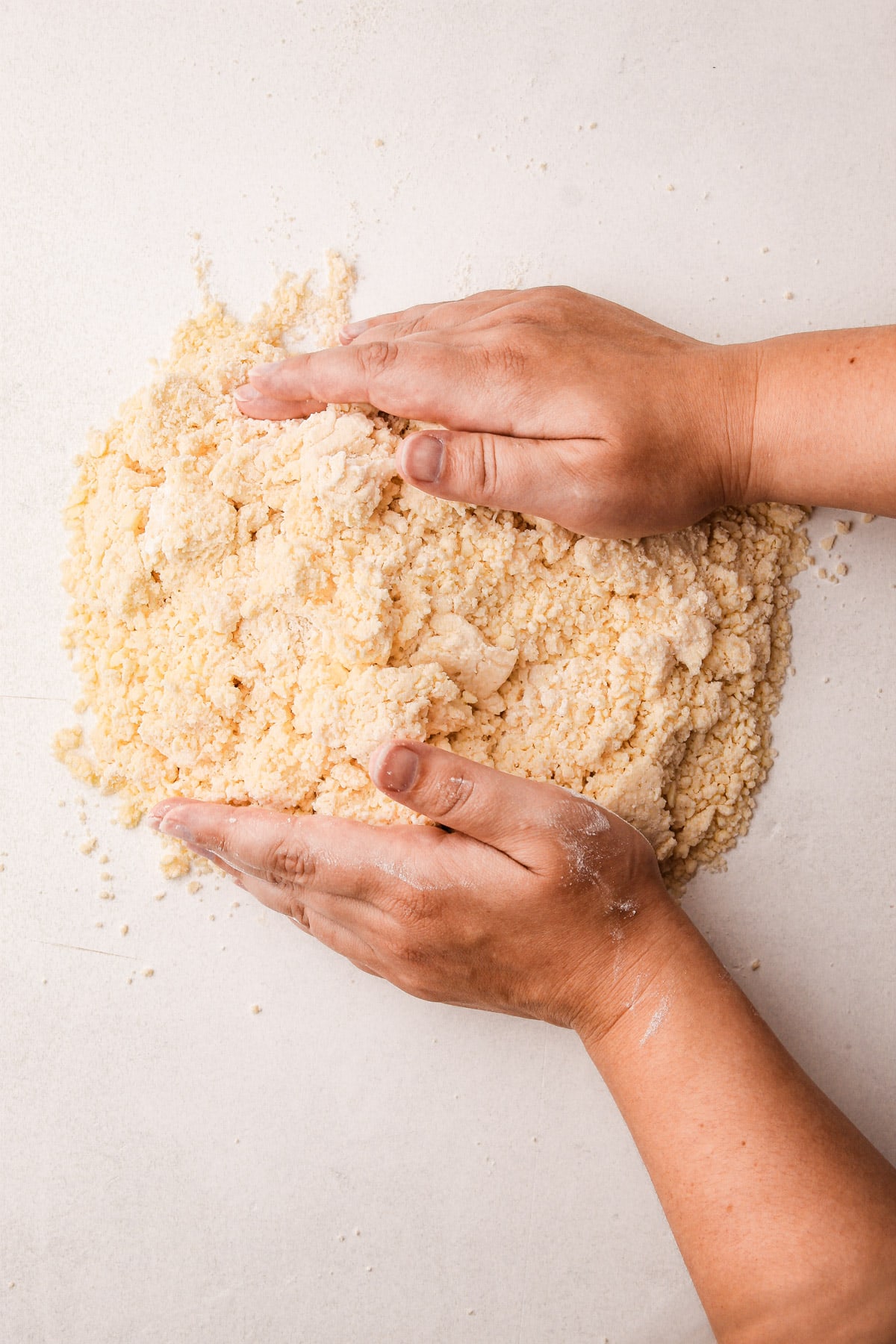 Two hands pushing together a crumbly pile of a flour and butter mixture on a counter surface