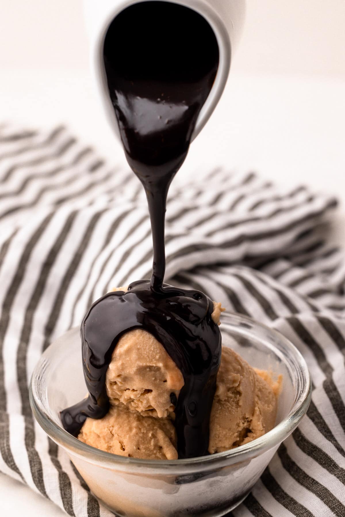 Hot fudge sauce pouring from a small white ceramic jug onto a bowl of peanut butter ice cream.