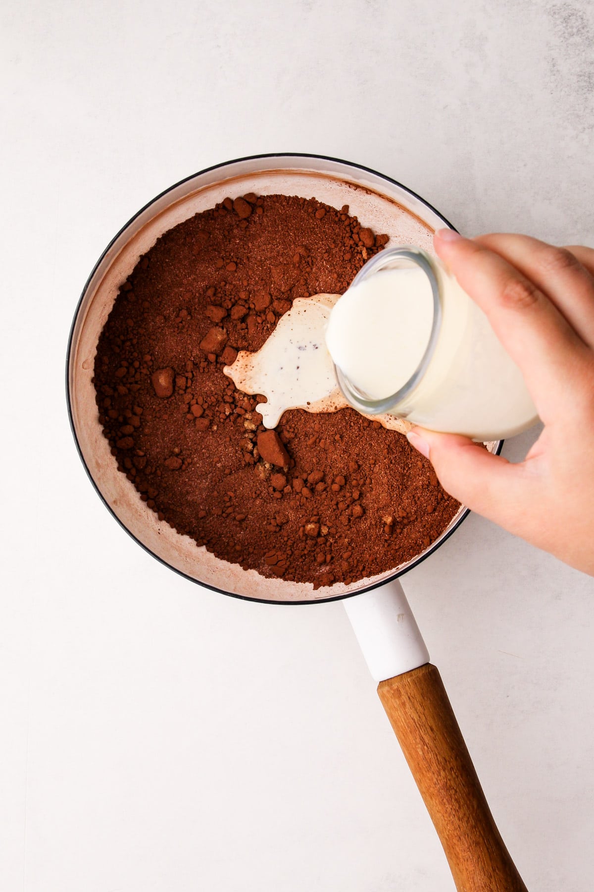 A pot with a cocoa mixture in it, with a hand pouring in cream to it from a glass jar