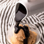 A hand pouring hot fudge sauce from a small white ceramic jug onto a bowl of peanut butter ice cream