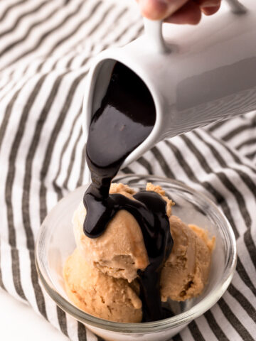 A hand pouring hot fudge sauce from a small white ceramic jug onto a bowl of peanut butter ice cream