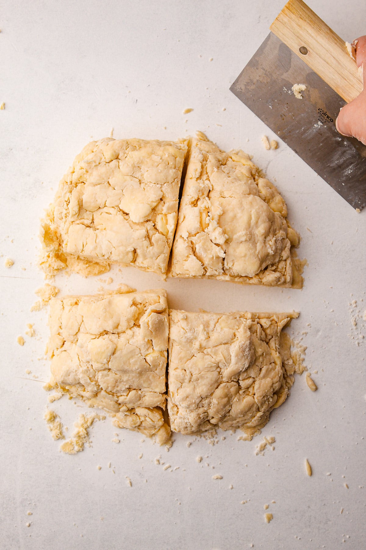 A hand holding a bench scraper over a mass of pie dough on a counter that has just been cut into quarters