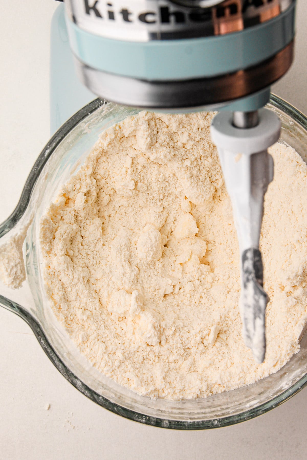 A sandy-looking flour mixture with pieces of butter in it in the bowl of a stand mixer, with the paddle attachment above the bowl