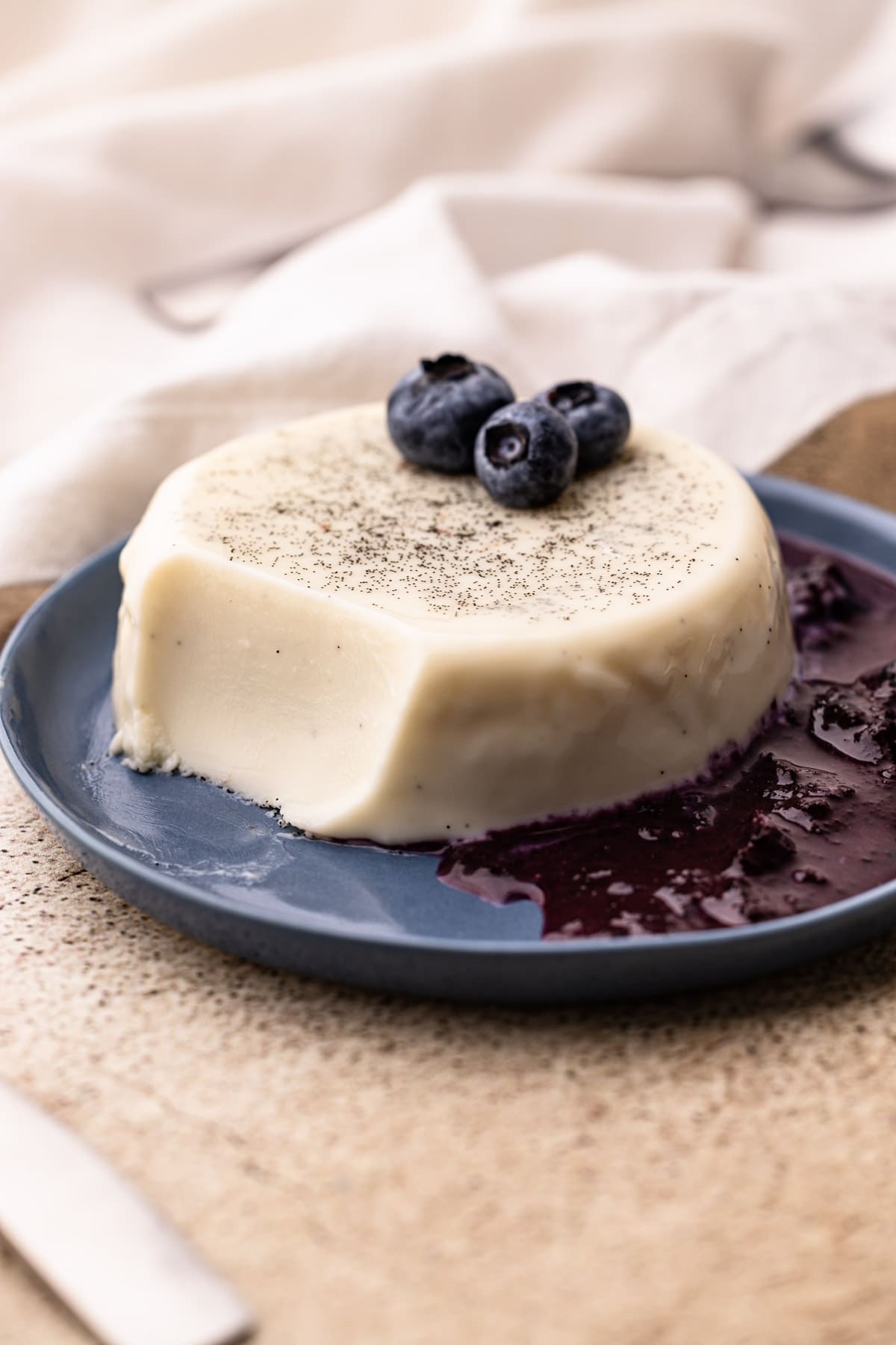 Panna cotta with a bite taken on a blue plate with berries and blueberry sauce.