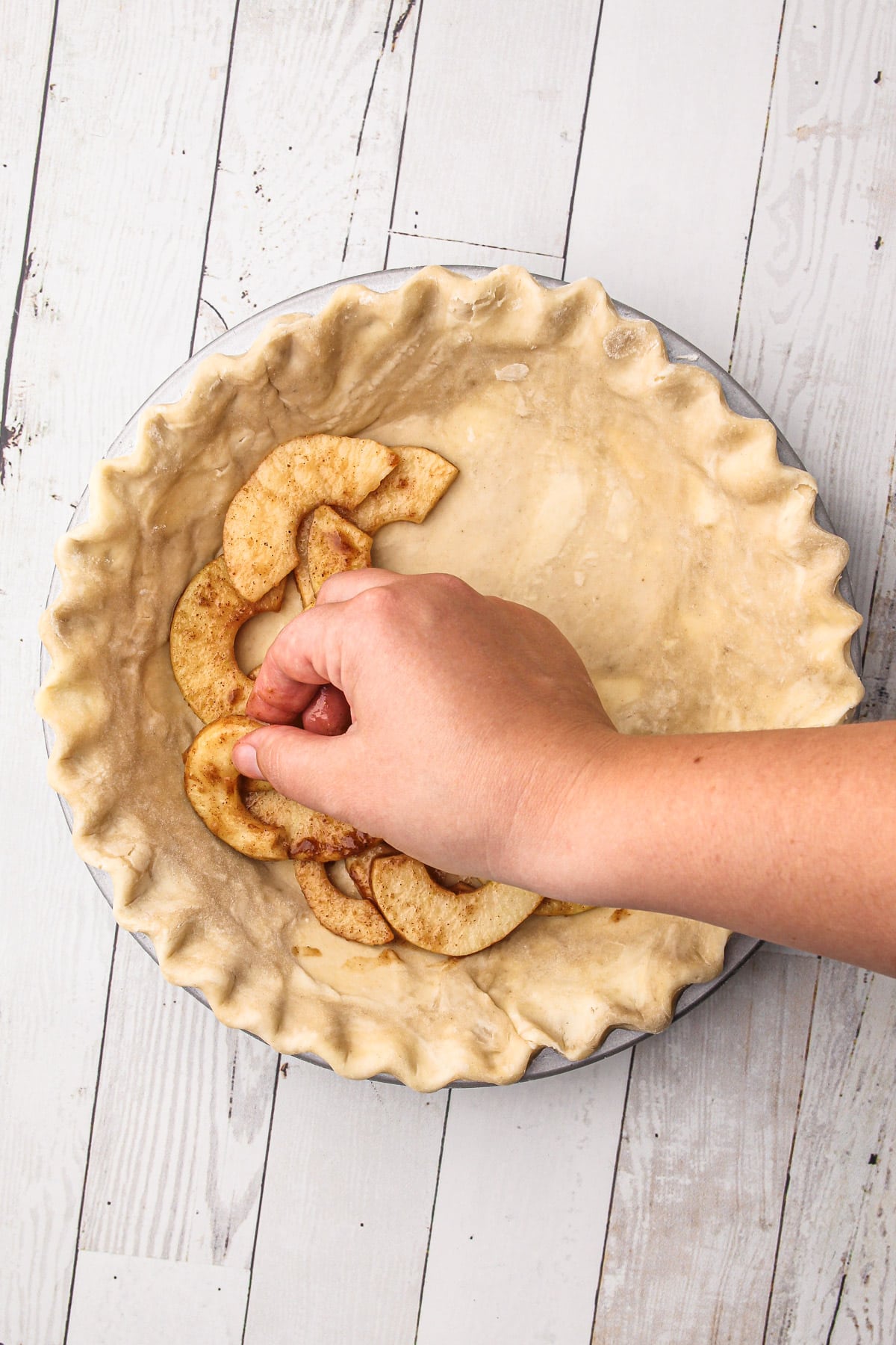 A hand placing slices of apple crumb pie filling into a shaped pie crust.