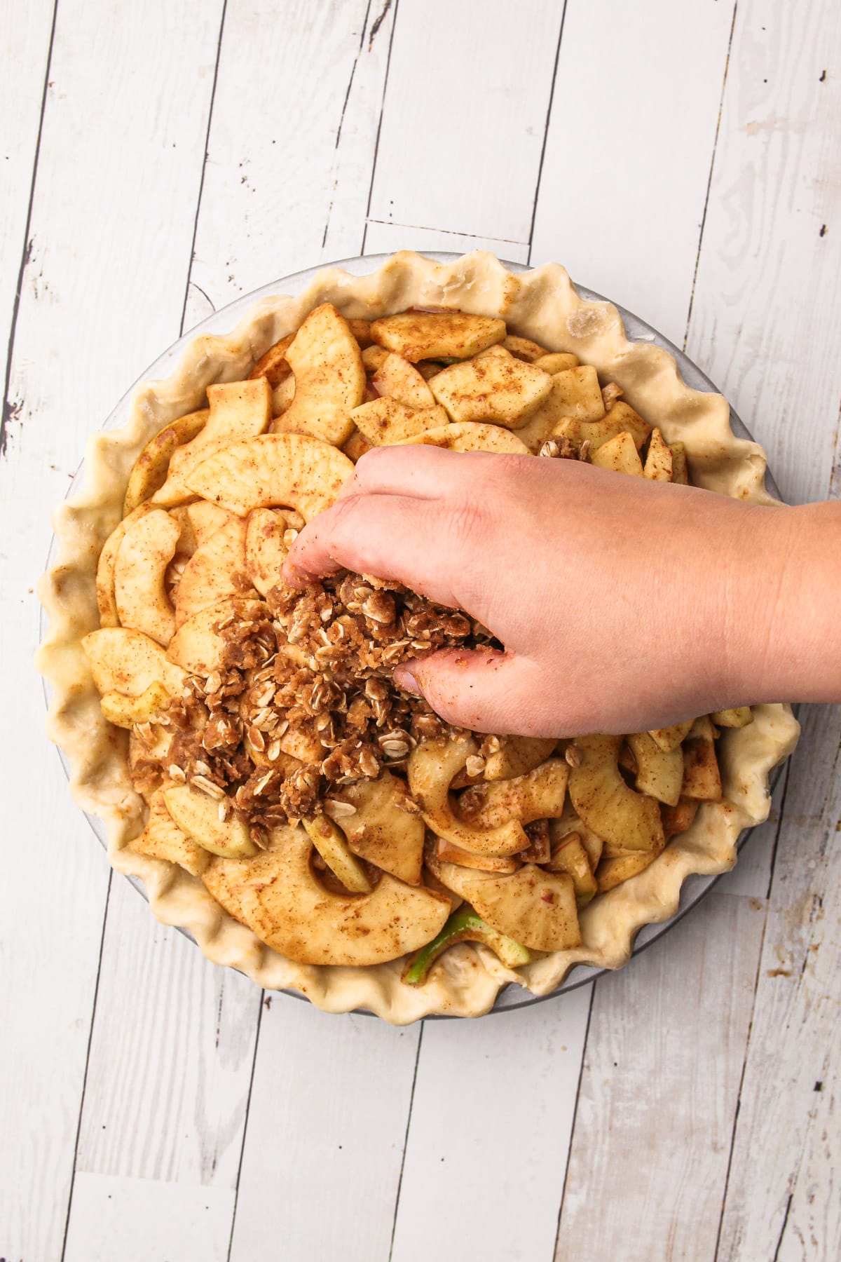 A hand sprinkling oat crumb topping onto an unbaked apple crumb pie.