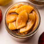 A jar filled to the top with apple pie filling surrounded by apples.