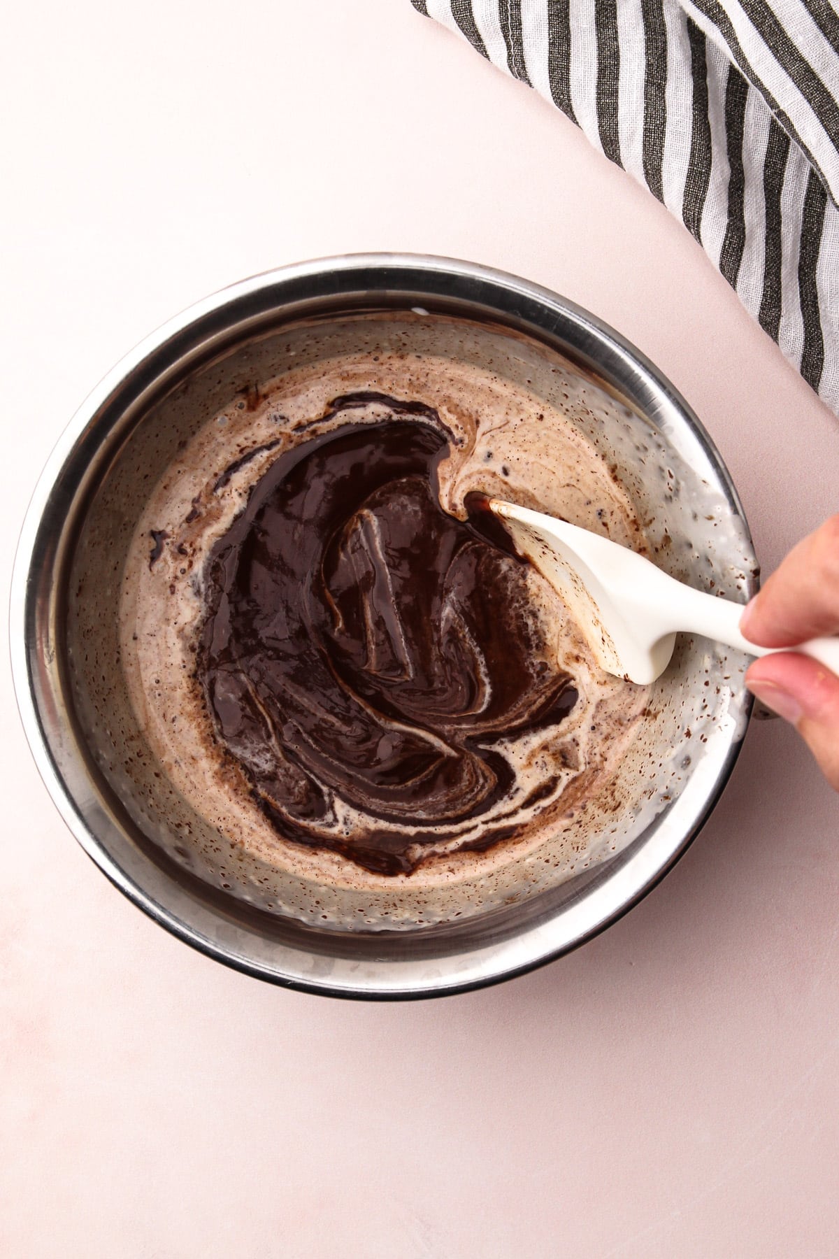 A hand stirring together melted chocolate and cream in a bowl with a rubber spatula to make chocolate ganache.