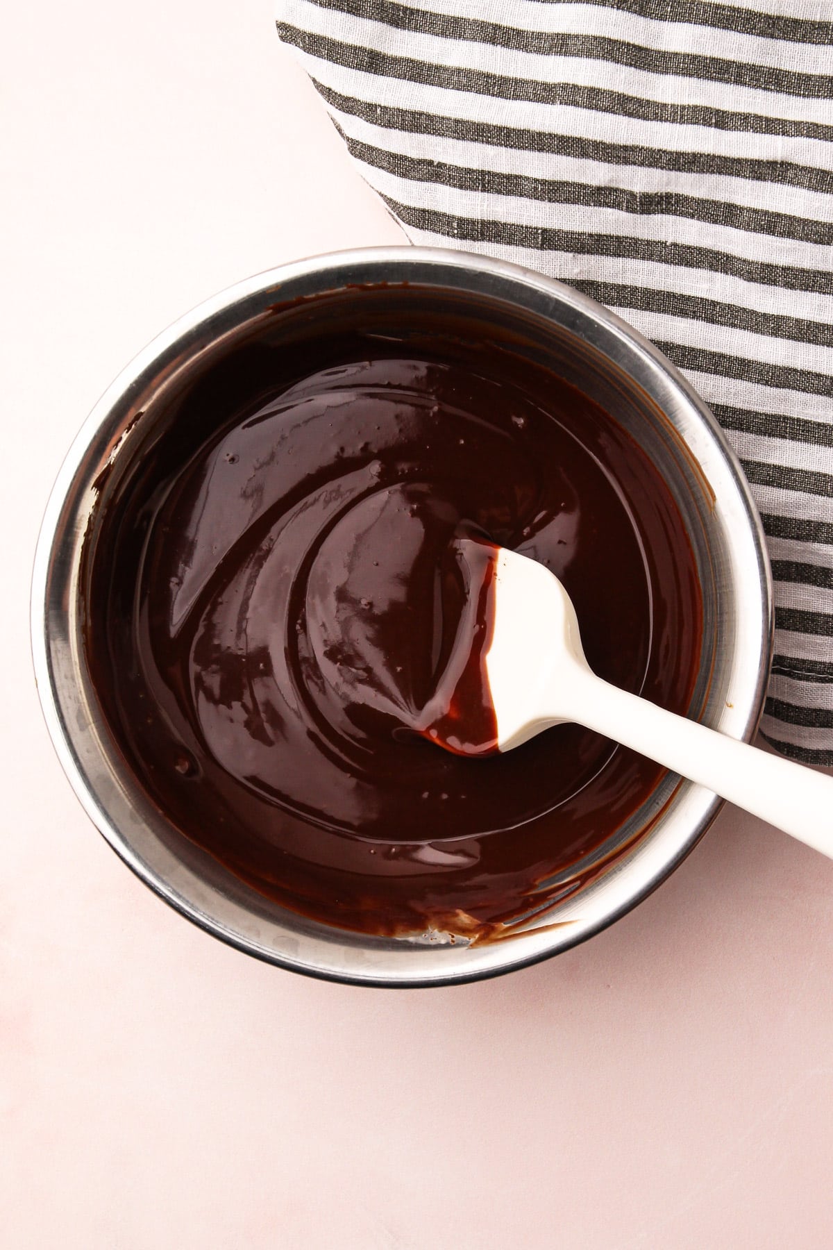 A rubber spatula stirring together a shiny, melted chocolate ganache sauce in a bowl.