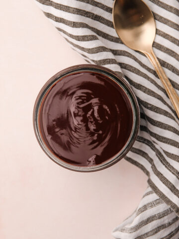 Shiny chocolate ganache sauce in a glass jar with a spoon next to it.