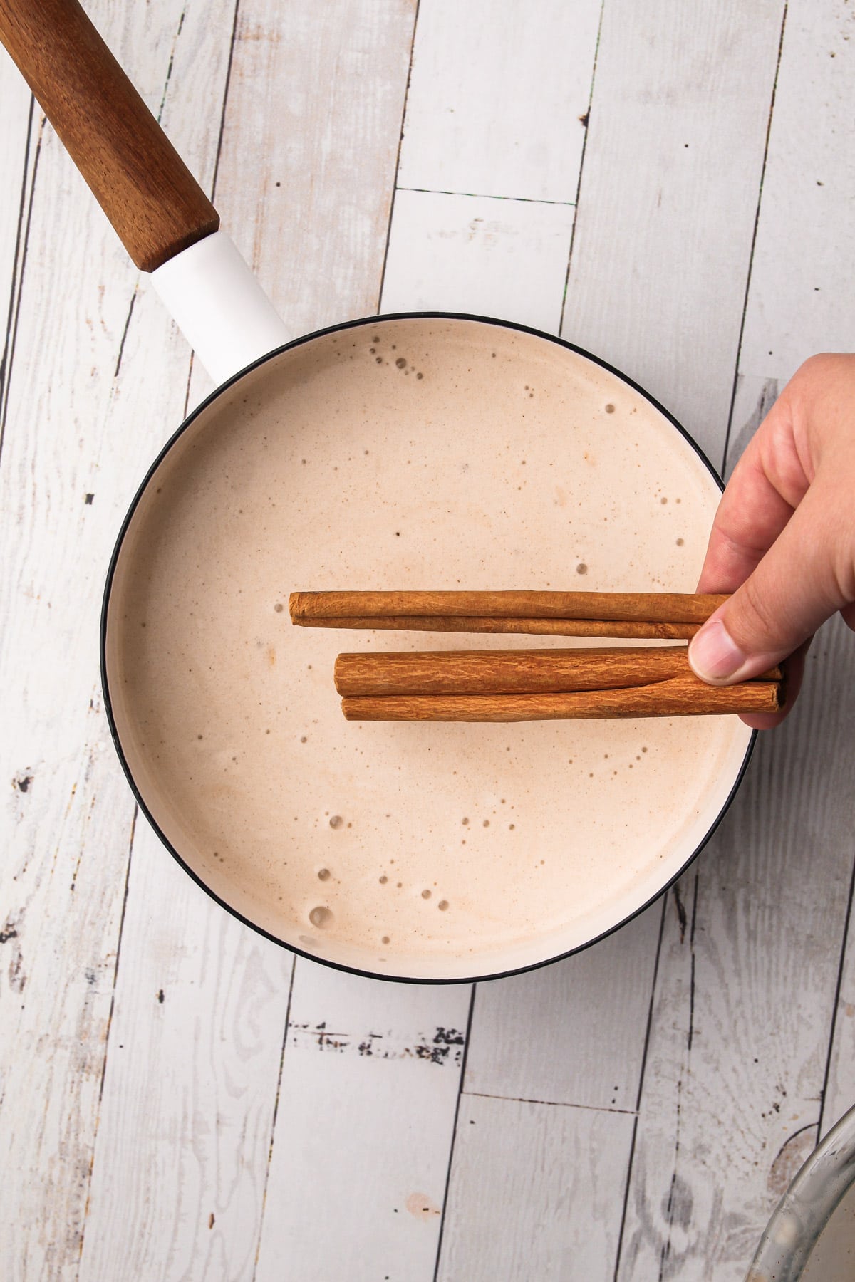 A hand adding two cinnamon sticks to a cinnamon ice cream base mixture in a saucepan.