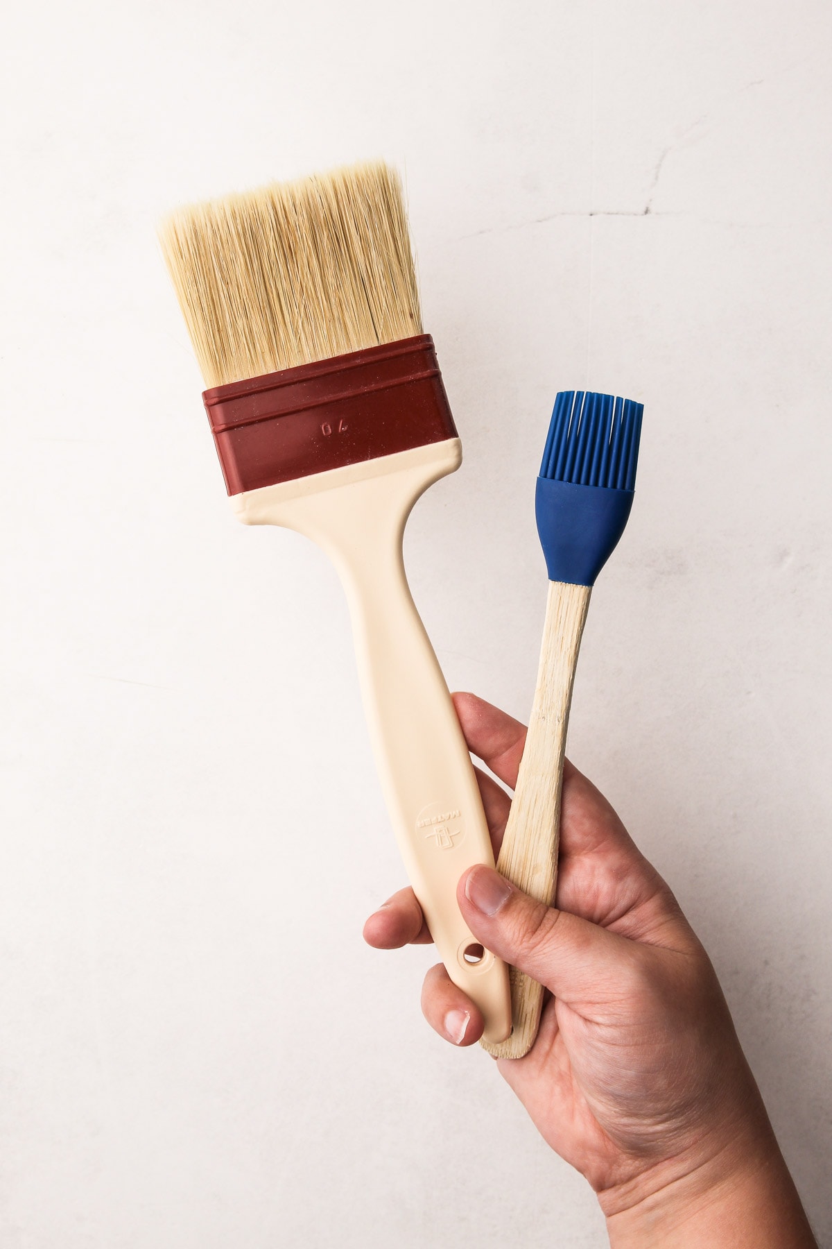 A hand holding a natural-bristle pastry brush and a silicone pastry brush, used as pie baking tools.