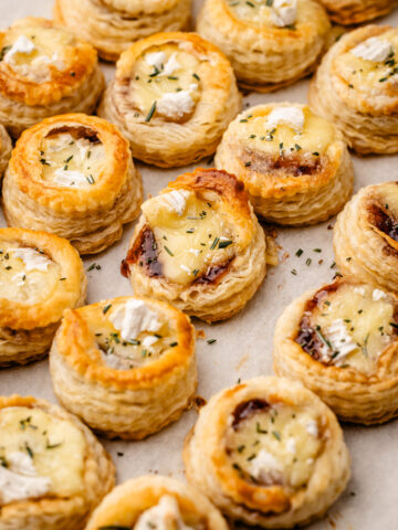 Many brie and jam puff pastry bites baked on a sheet pan with a sprinkle of rosemary.