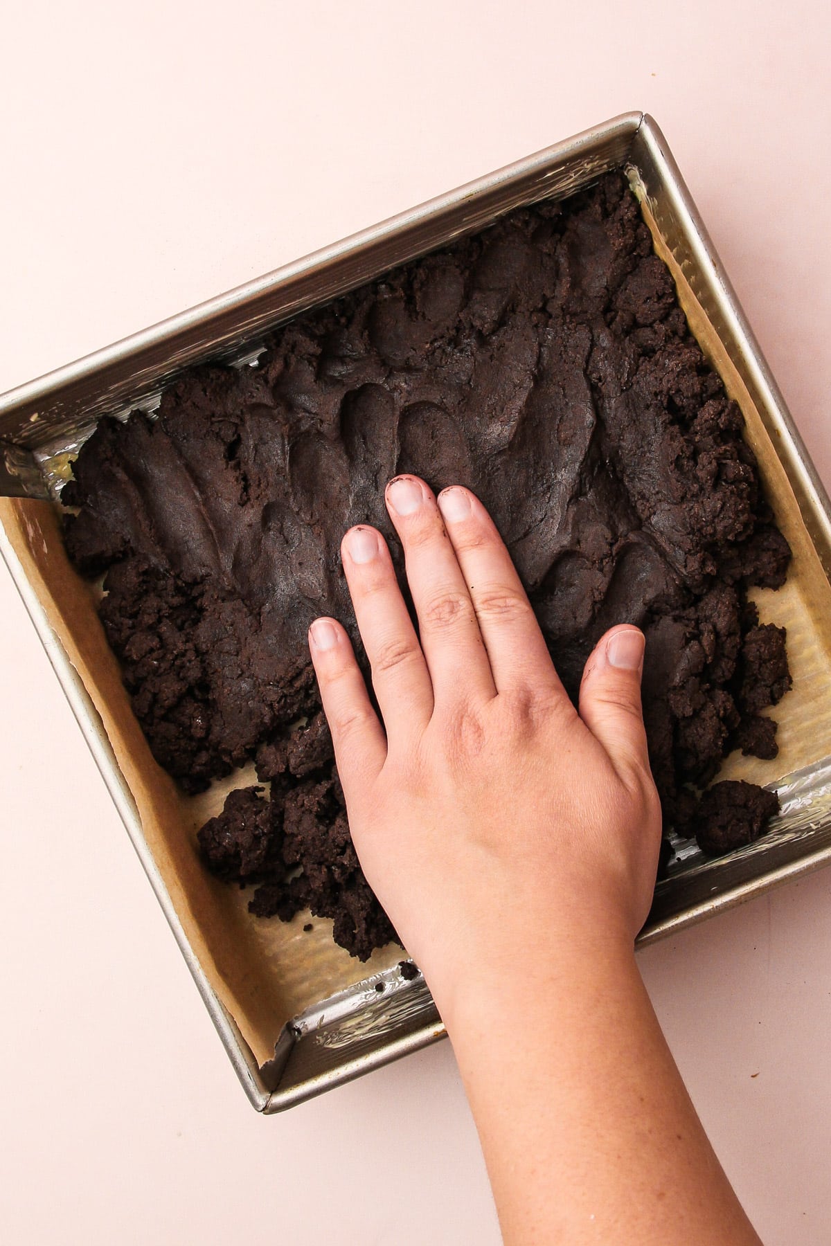 A hand pressing dough for chocolate shortbread cookies into a square baking pan.