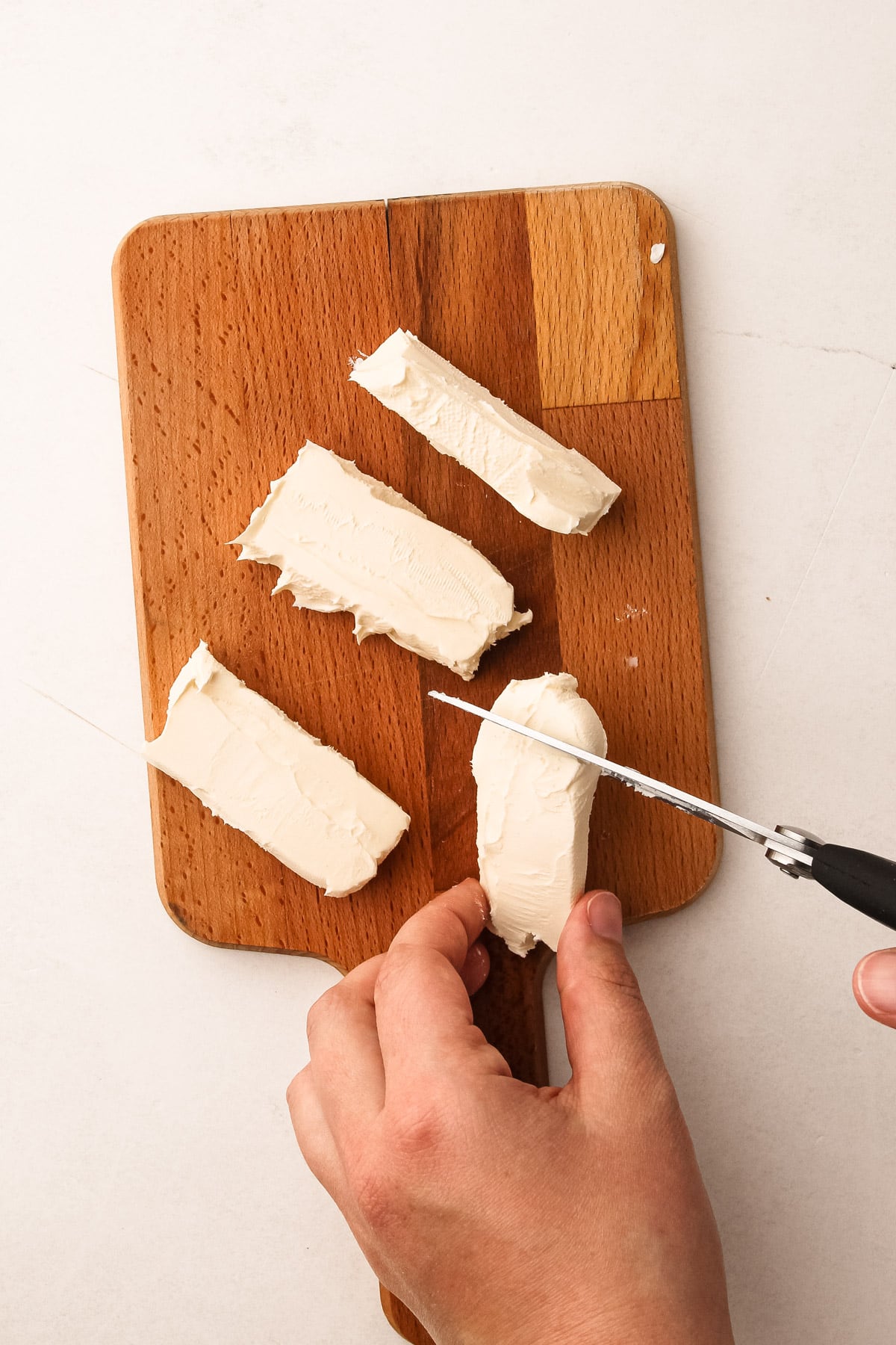 A hand using kitchen shears to cut cream cheese into pieces for puff pastry pepper jelly bites.