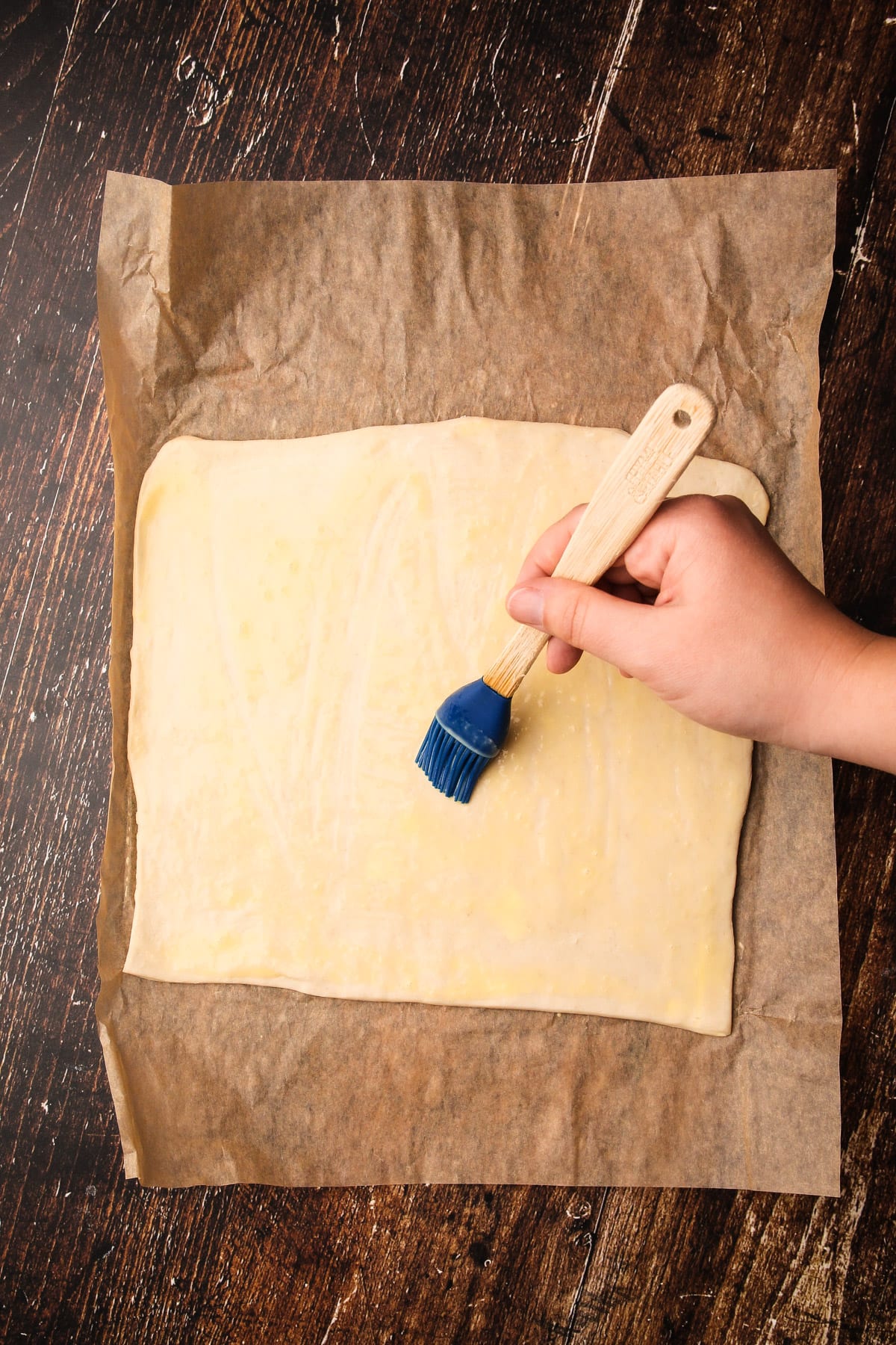 A hand using a pastry brush to paint a square of pastry with egg wash for puff pastry cheese straws.