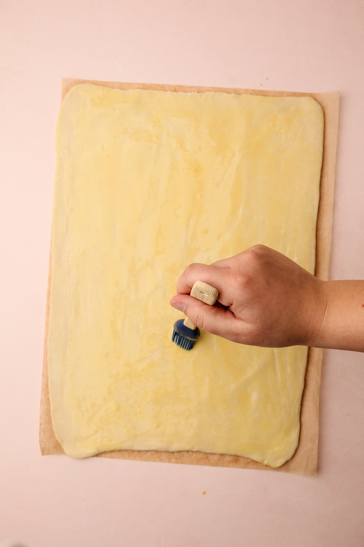 A hand using a pastry brush to brush a sheet of puff pastry with egg wash.