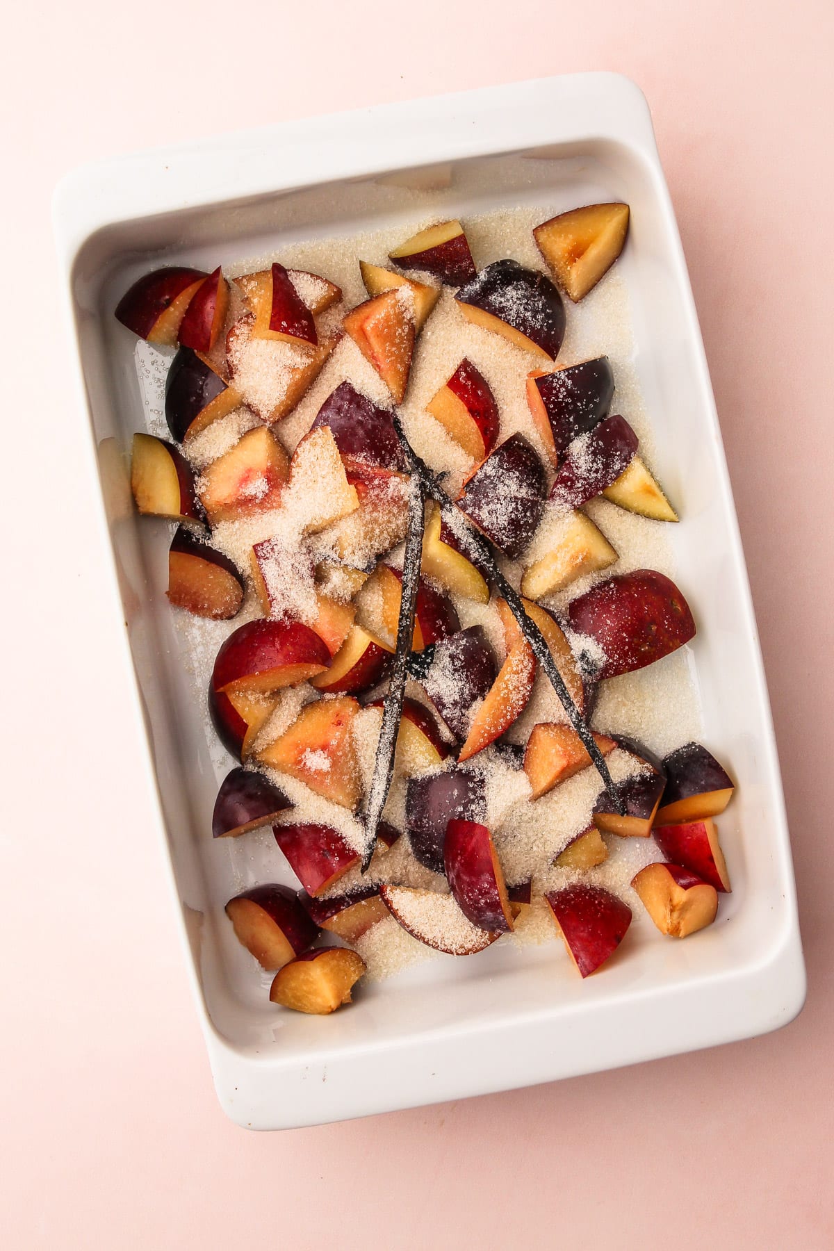 A baking dish filled with chunks of raw plums, sugar, and vanilla beans for roasted plum pie.