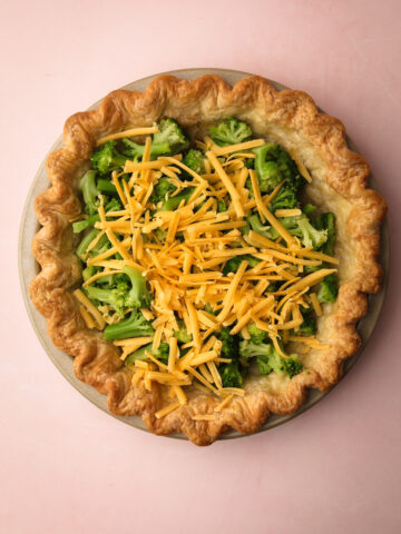 A baked pie shell filled with broccoli florets and shredded cheddar cheese for Broccoli Cheddar Quiche.