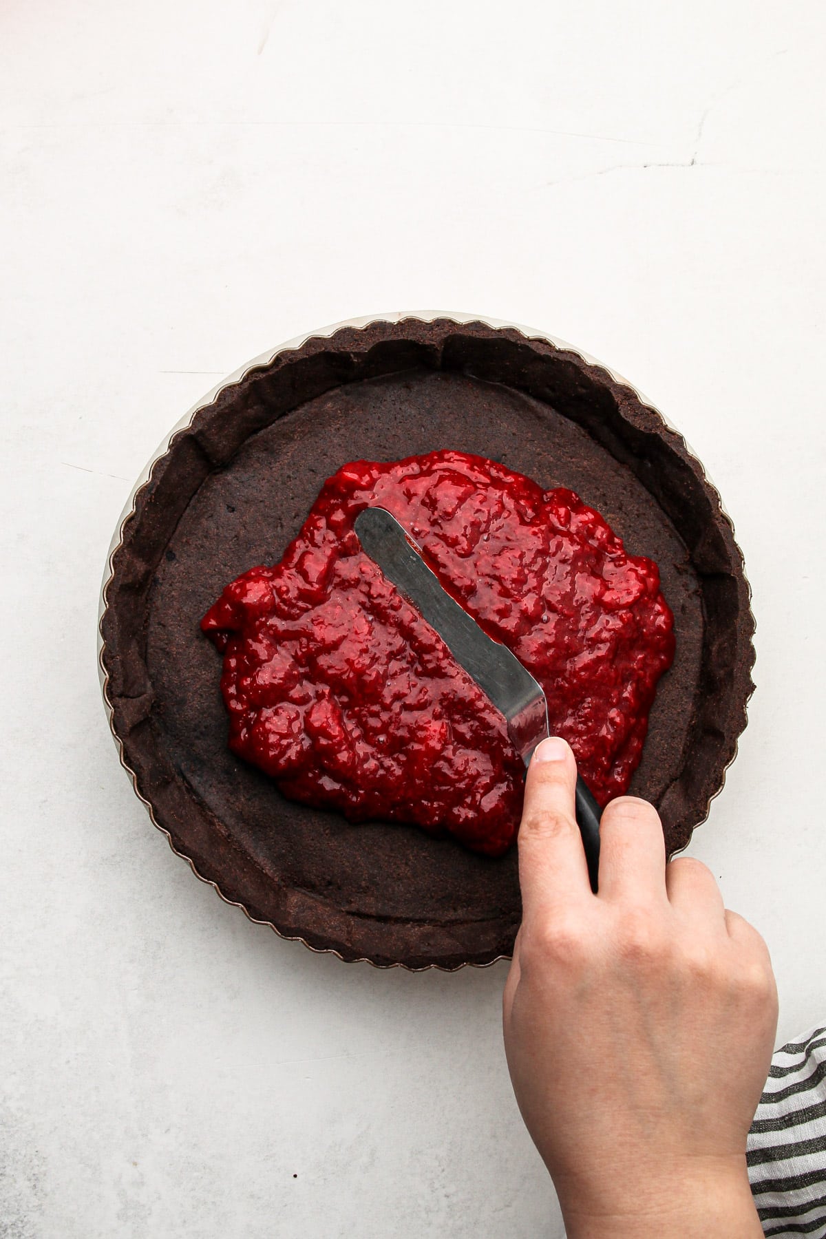 A hand using an offset spatula to spread strawberry filing into a chocolate tart shell for chocolate strawberry pie.