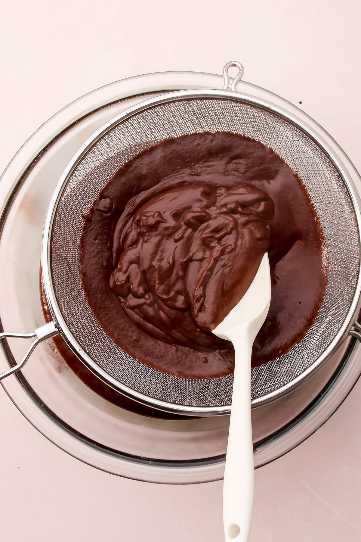 Cooked custard for dark chocolate pudding being pushed with a spatula through a fine-mesh strainer into a bowl.