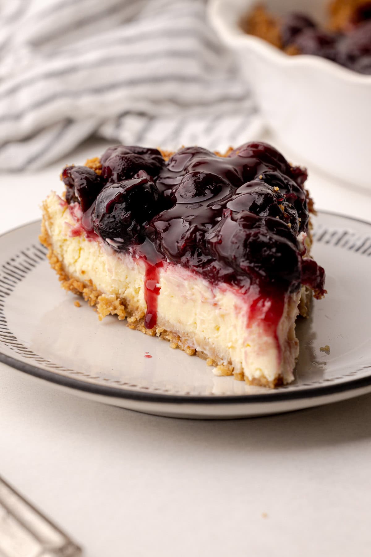 A slice of cherry cheesecake pie on a plate with a bit of juicy cherry dripping down.