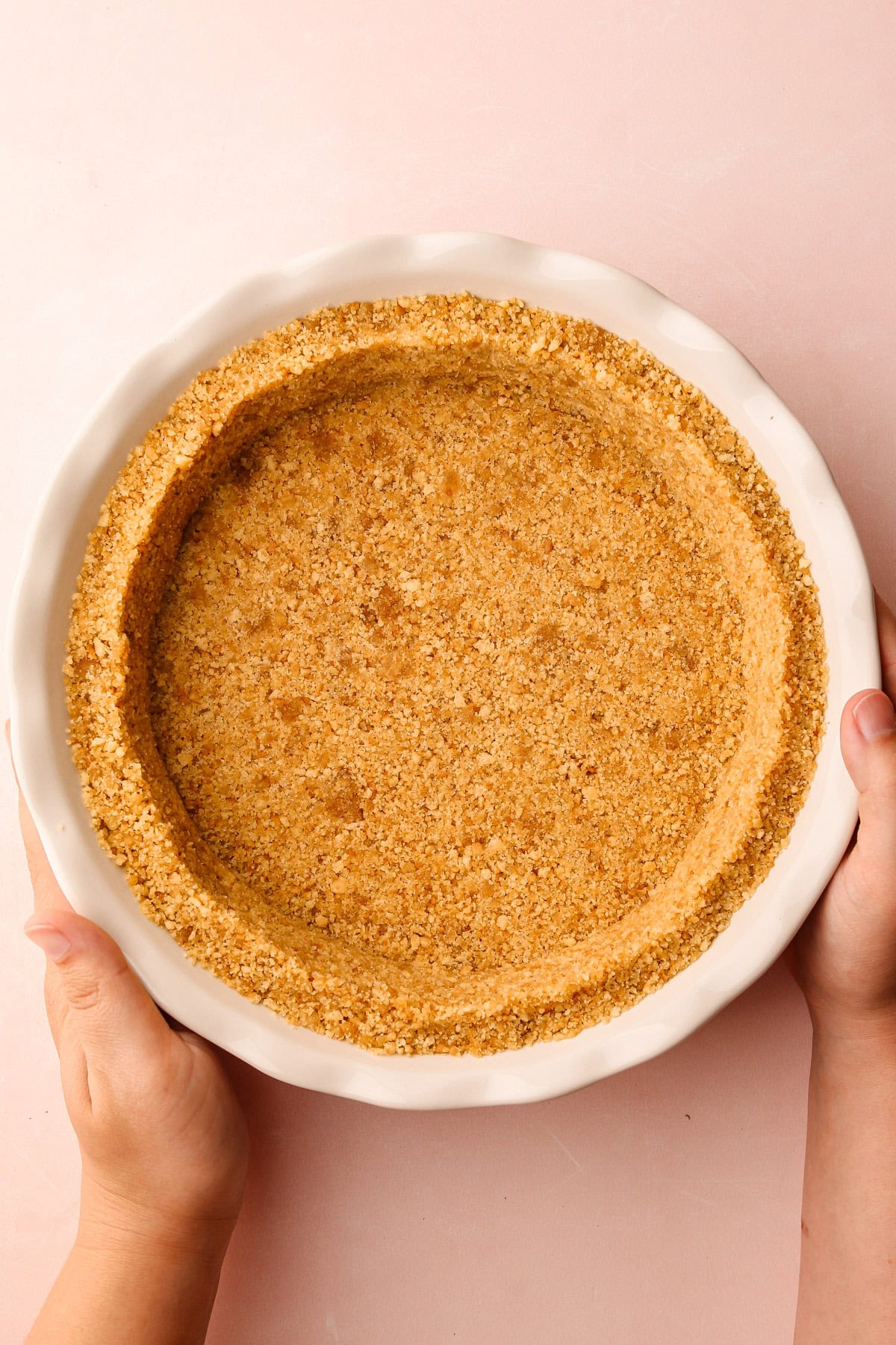 Hands holding a vanilla wafer crumb crust in a white pie plate.