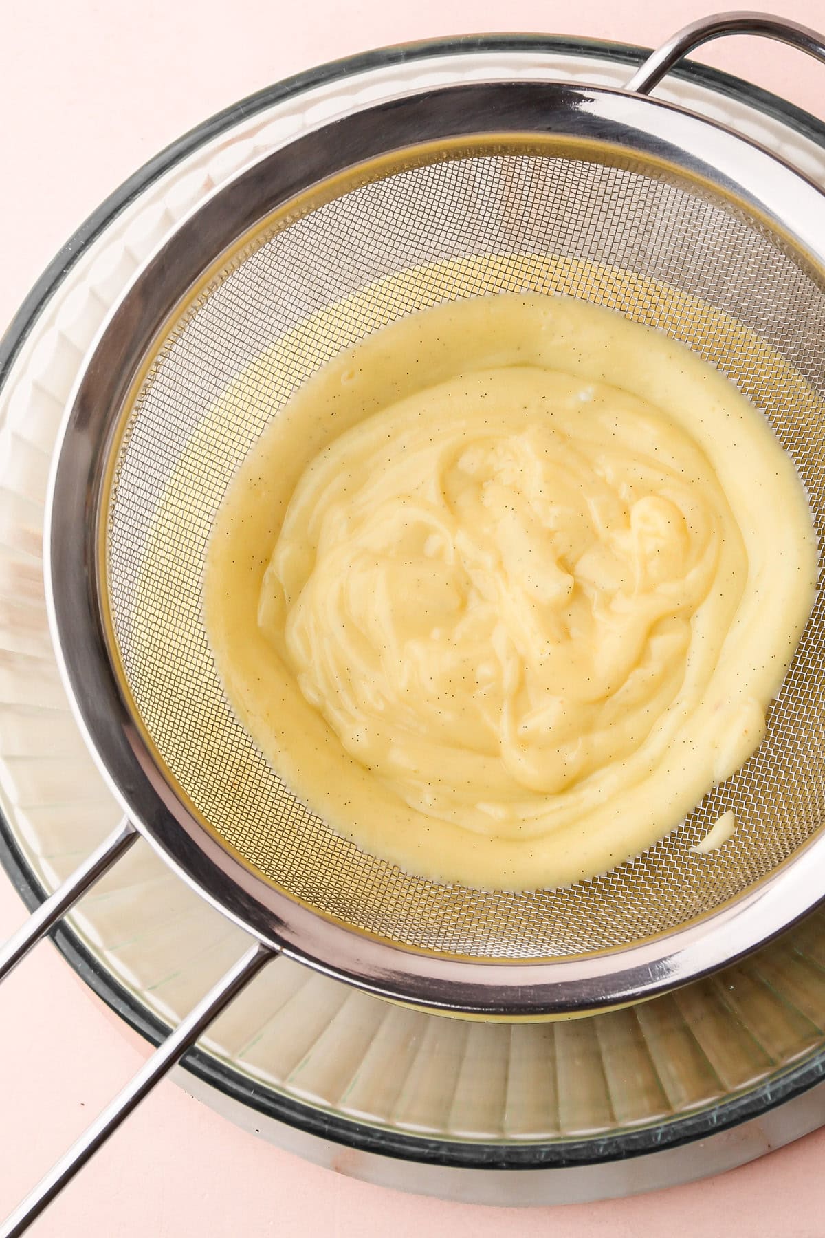 Thickened pastry cream being strained through a fine mesh sieve into a glass bowl.