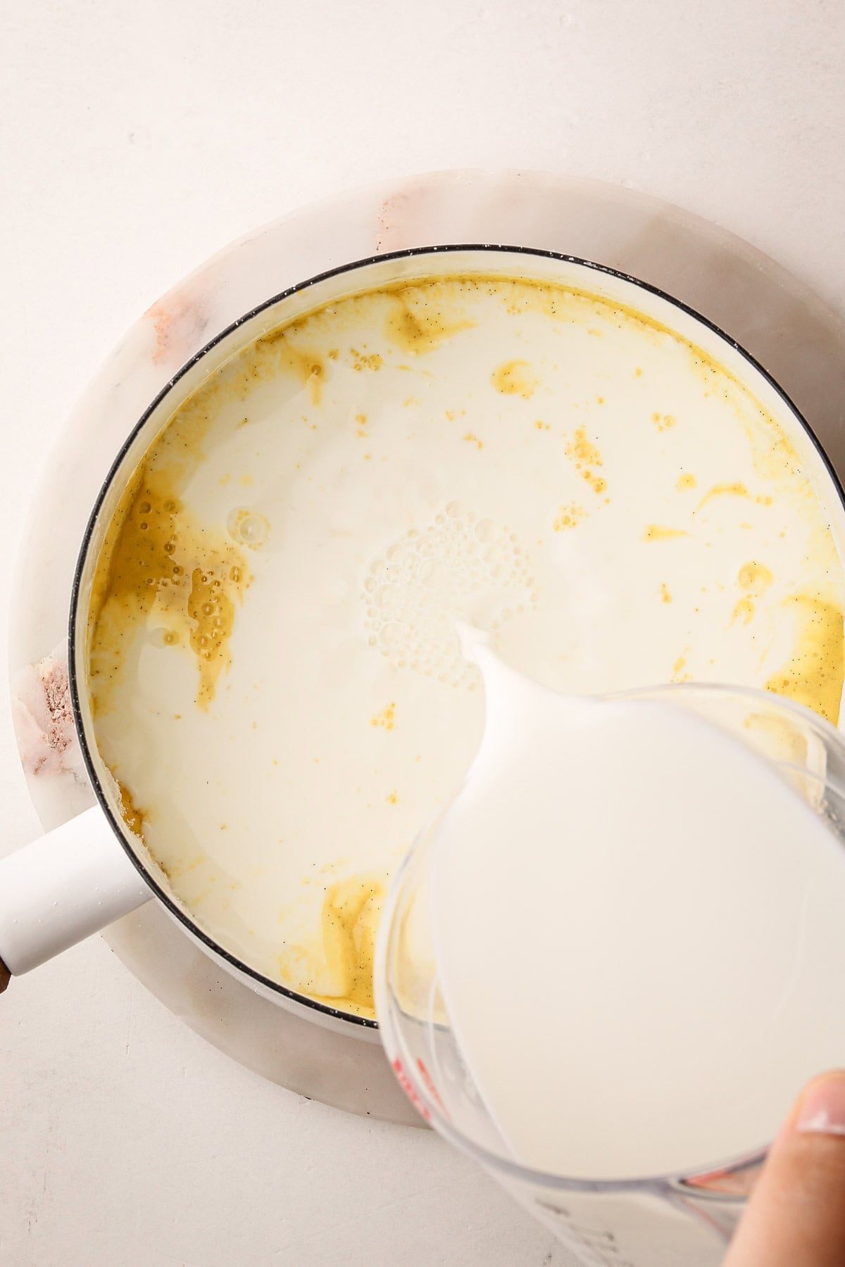 Milk being poured into a white saucepan containing a yellow mixture of eggs and vanilla bean seeds, to start the pastry cream.