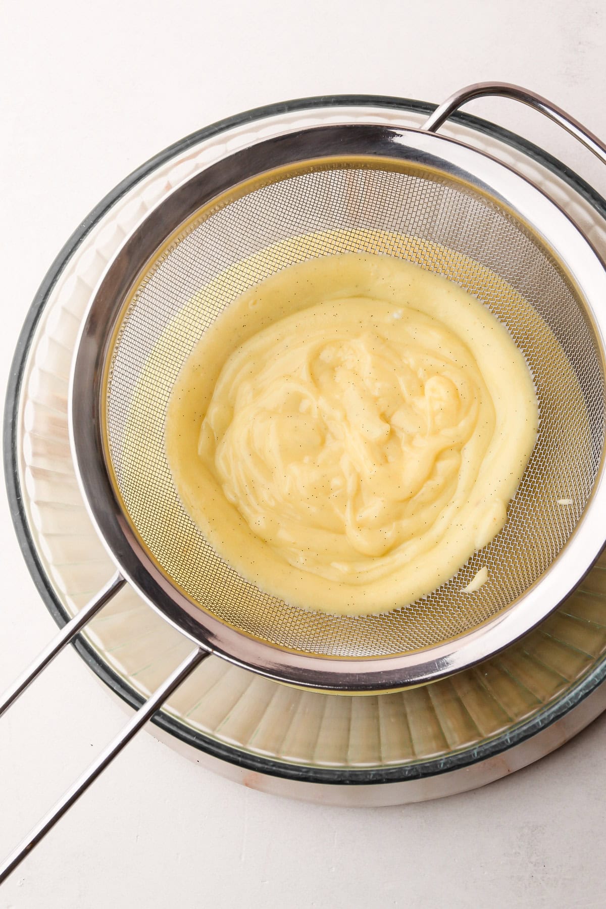 Thickened pastry cream being strained through a fine mesh sieve into a glass bowl.