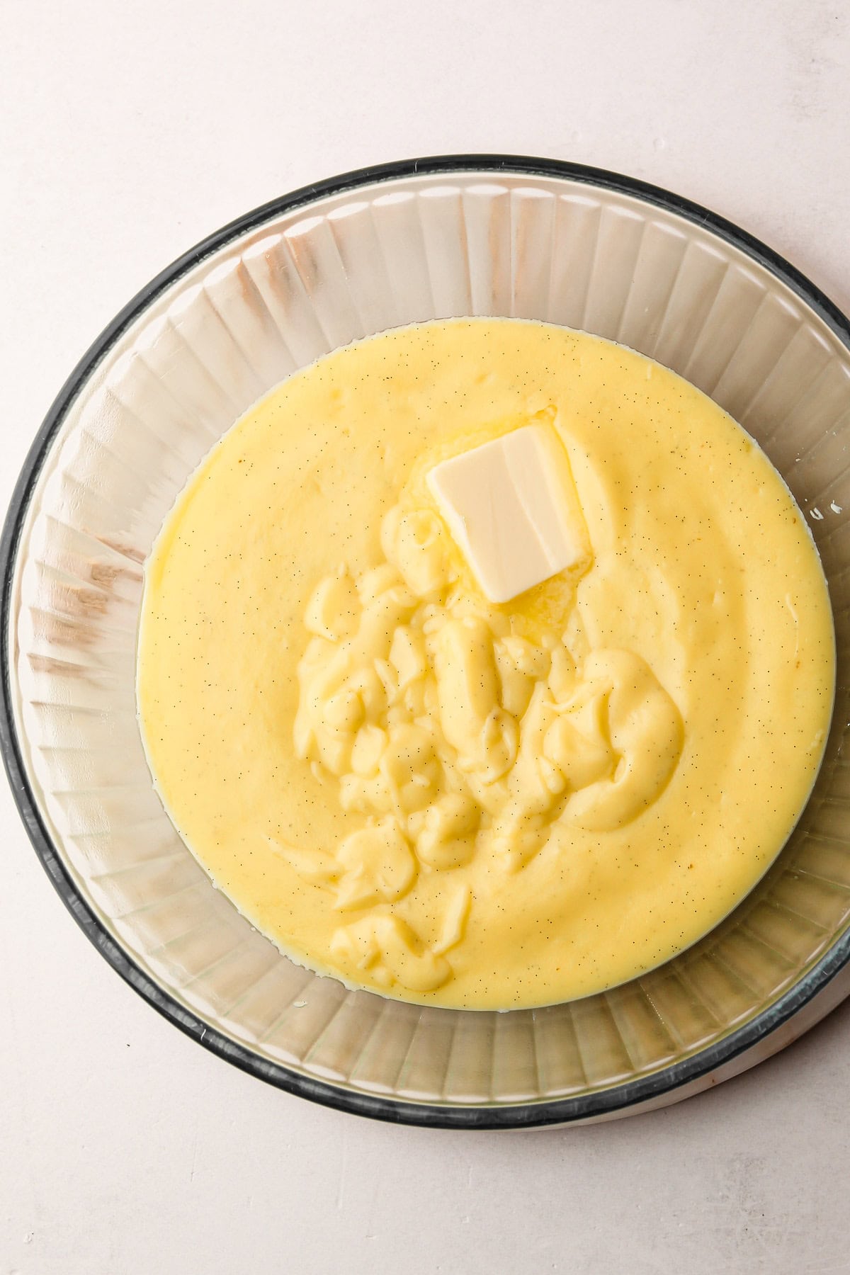 A pat of butter placed on top of freshly made pastry cream recipe in a glass bowl.