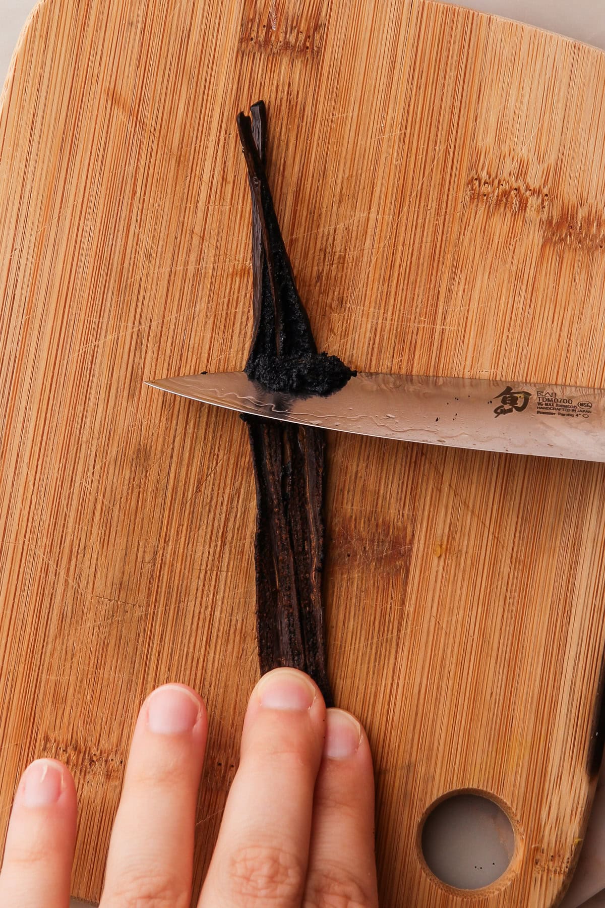 Close-up of a knife slicing through a vanilla bean pod, revealing the seeds inside, on a small wooden cutting board.