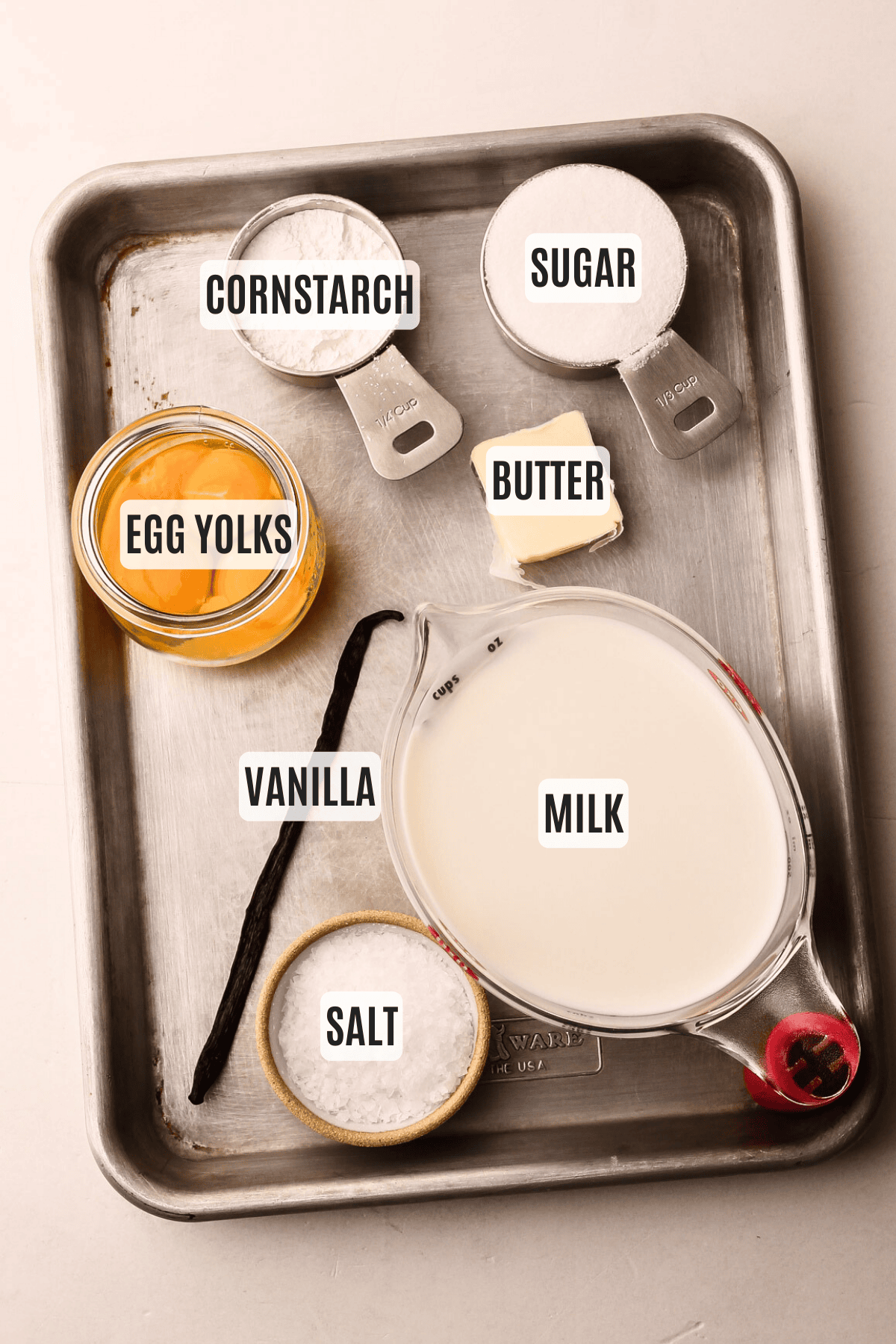 Various ingredients for pastry cream labeled: cornstarch, sugar, butter, egg yolks, vanilla, milk, and salt, displayed on a metal tray.