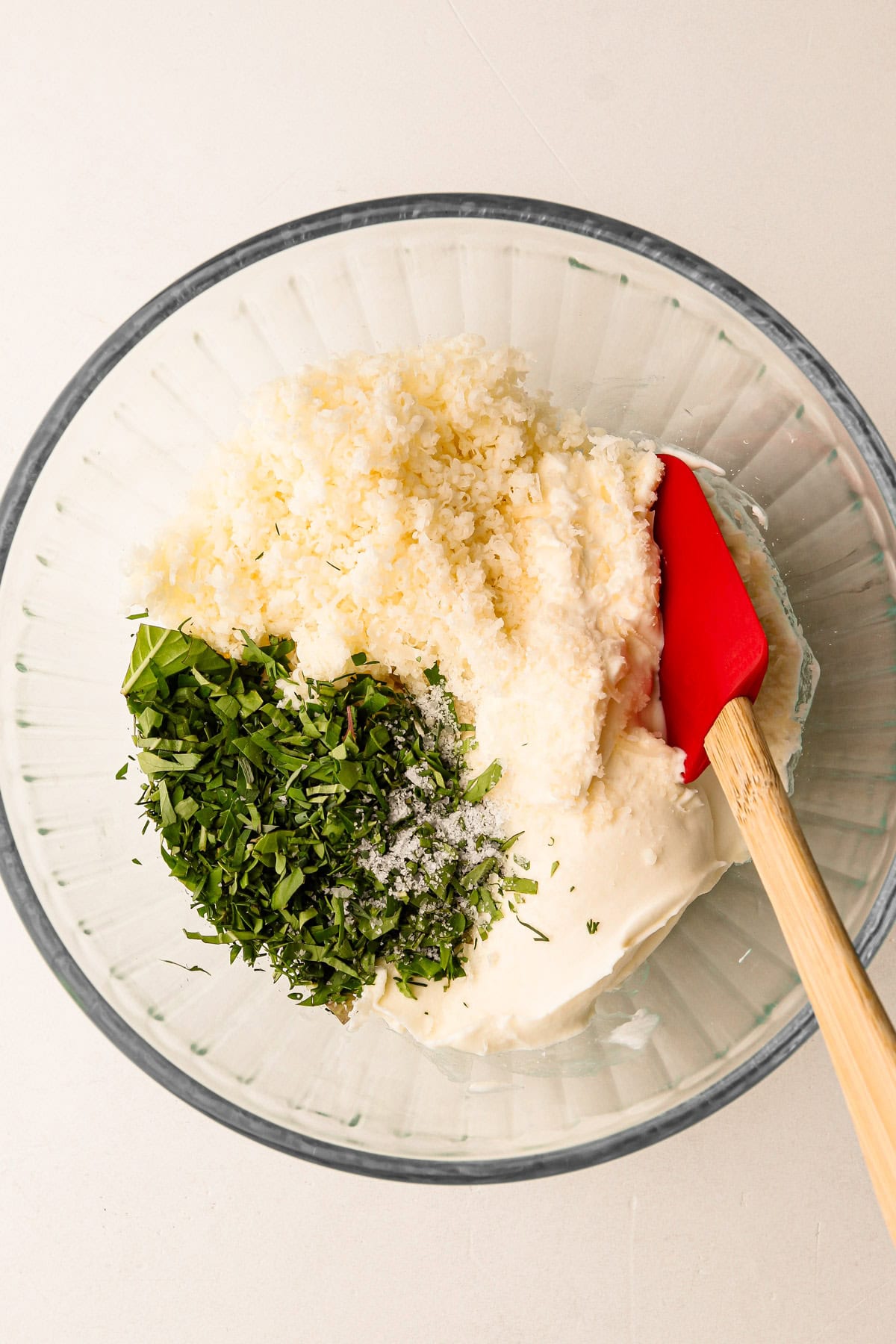 Crème fraîche, herbs, and cheese mixture in a glass bowl with a red spatula.