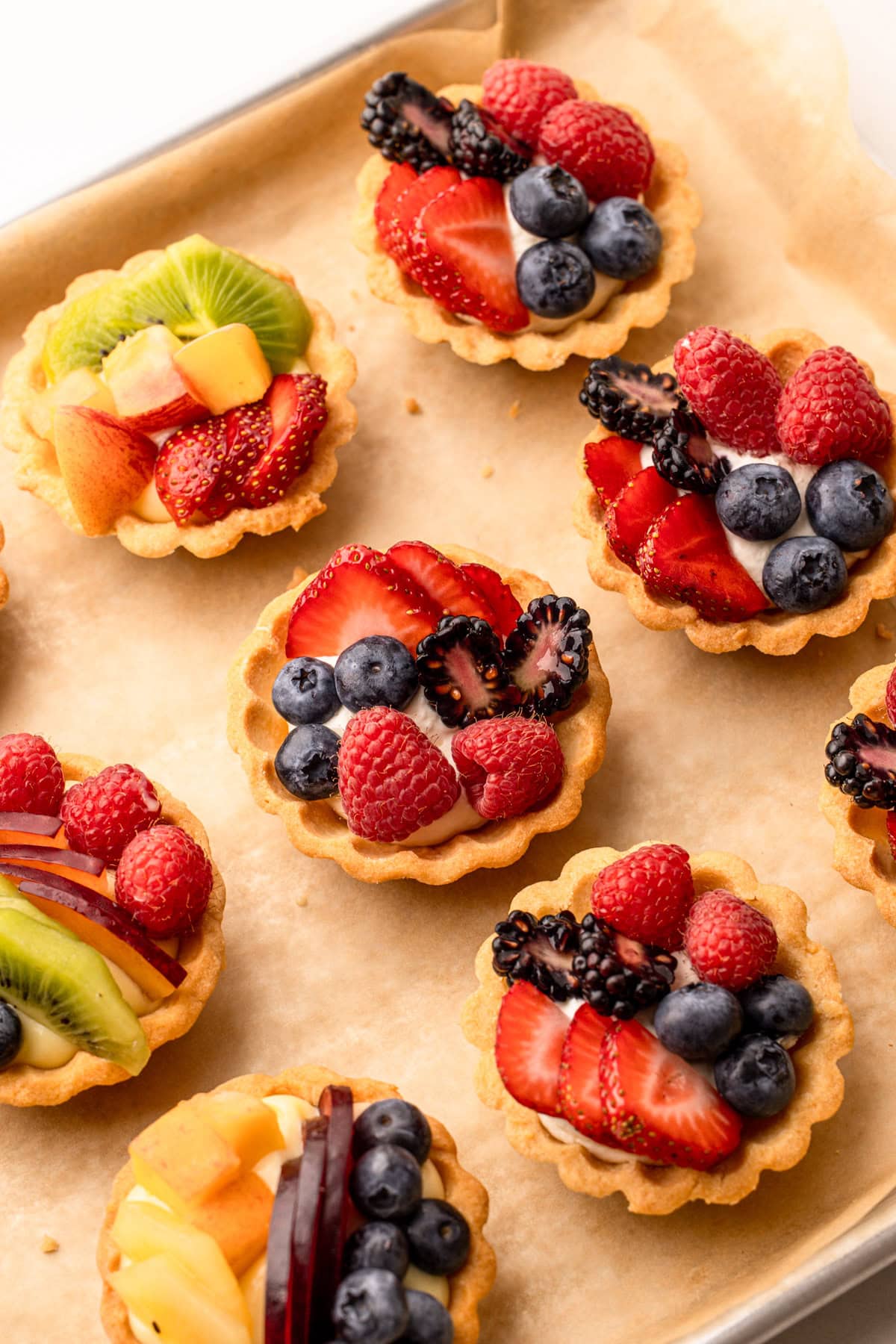 Several mini fruit tarts with mixed berries and kiwi on a baking sheet.