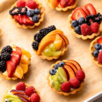 Several mini fruit tarts with mixed berries and kiwi on a baking sheet.