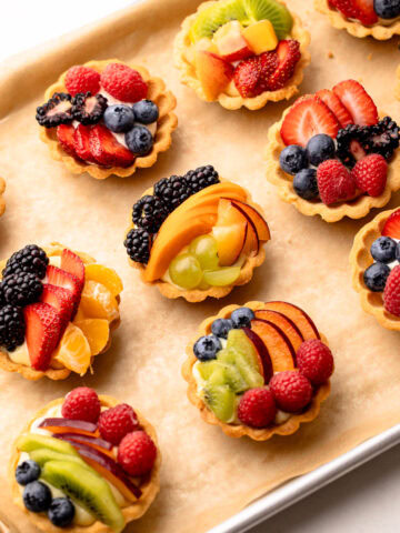 Several mini fruit tarts with mixed berries and kiwi on a baking sheet.